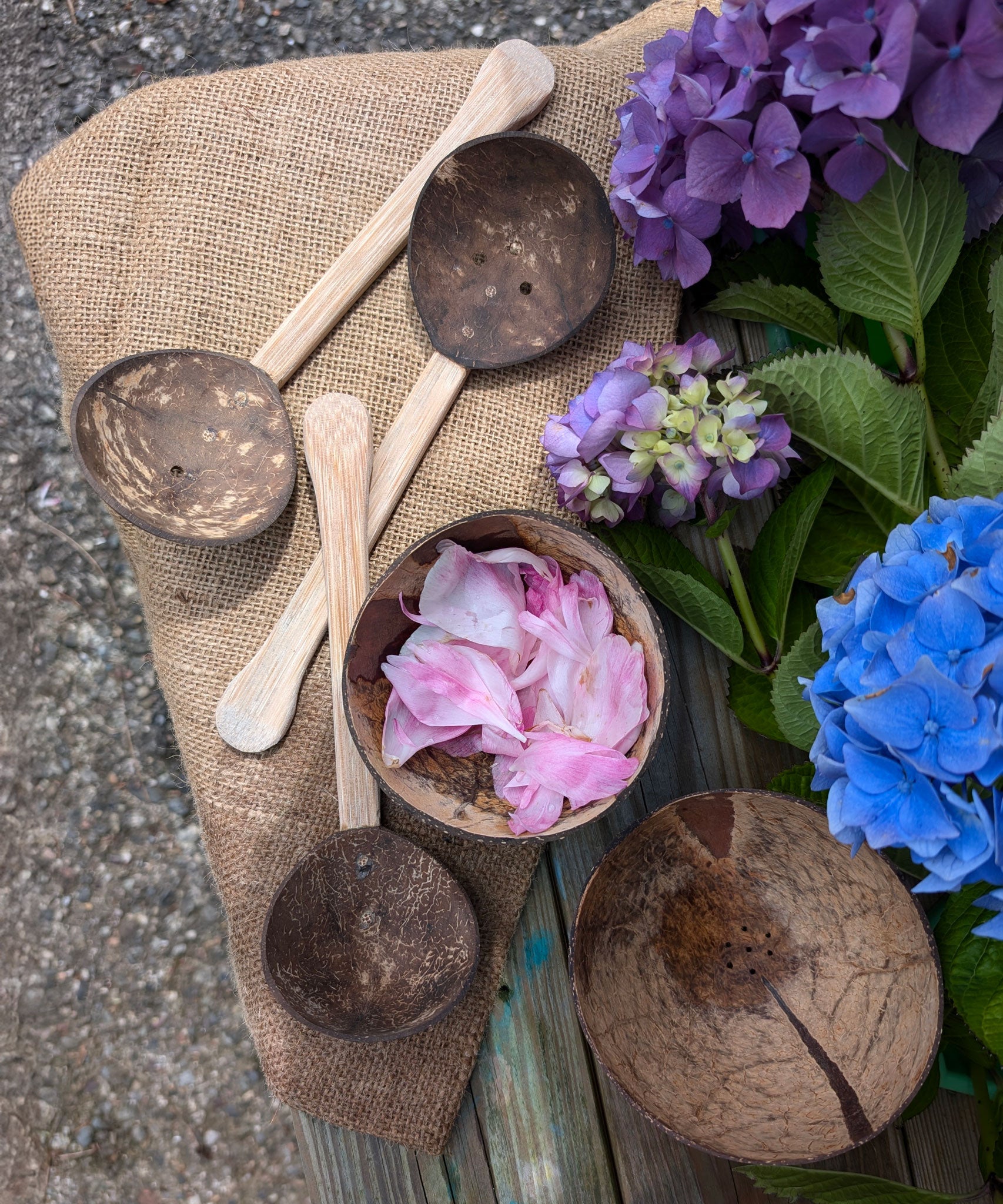 The utensils and bowls Lanka Kade Coconut Shell Water Playset placed on a jute material. There are pink coloured peony petal in the coconut bowl. Blue and purple coloured hydrangeas can be seen to the side. 