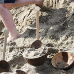Lanka Kade coconut bowls and utensils placed on sand on a beach. A child can be seen running past in the background with bits of sand in the air as he goes past.