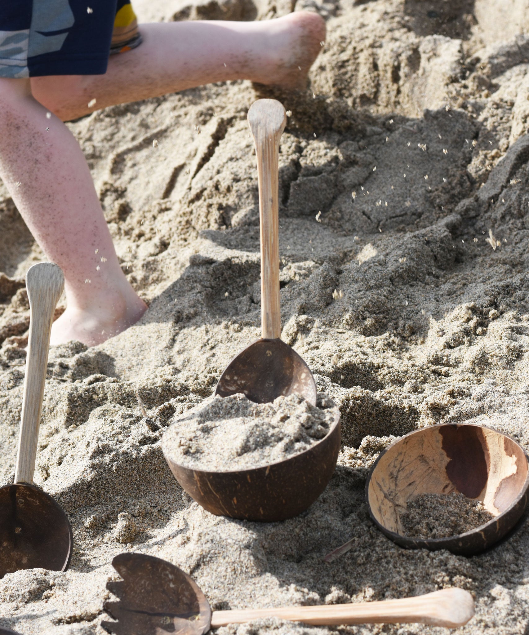 Lanka Kade coconut bowls and utensils placed on sand on a beach. A child can be seen running past in the background with bits of sand in the air as he goes past.