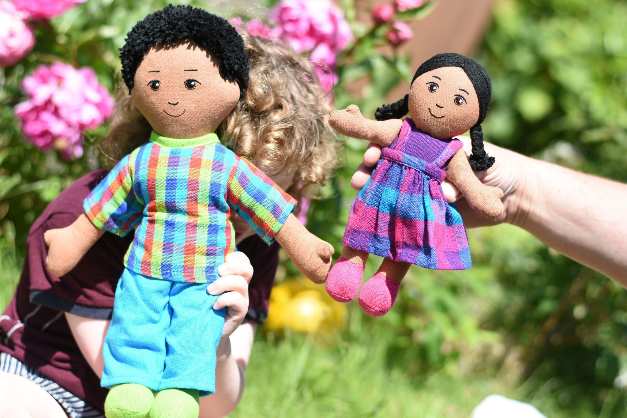 A close up of a child holding up a Lanka Kade dad doll with brown skin and black hair next to an adult holding up a girl doll with brown skin and black hair. 