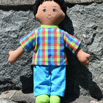 A Lanka Kade dad doll with brown skin and black hair leaning on a stone wall outdoors. 