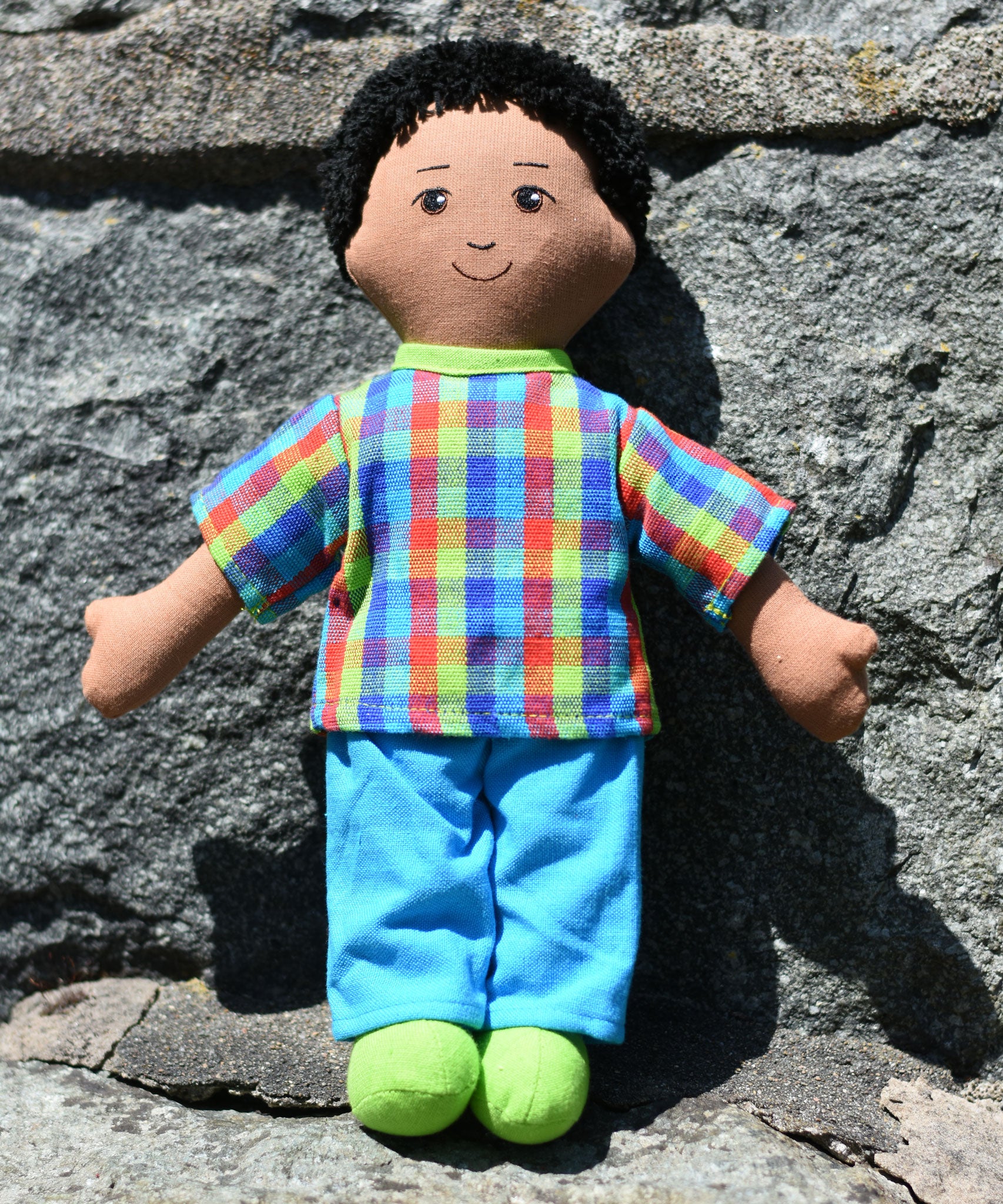 A Lanka Kade dad doll with brown skin and black hair leaning on a stone wall outdoors. 