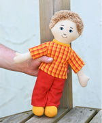 A Fair Trade Lanka Kade Dad doll shown in an adult's hand. The doll has white skin and white hair and is wearing a t-shirt and trousers made from sari material offcuts. 