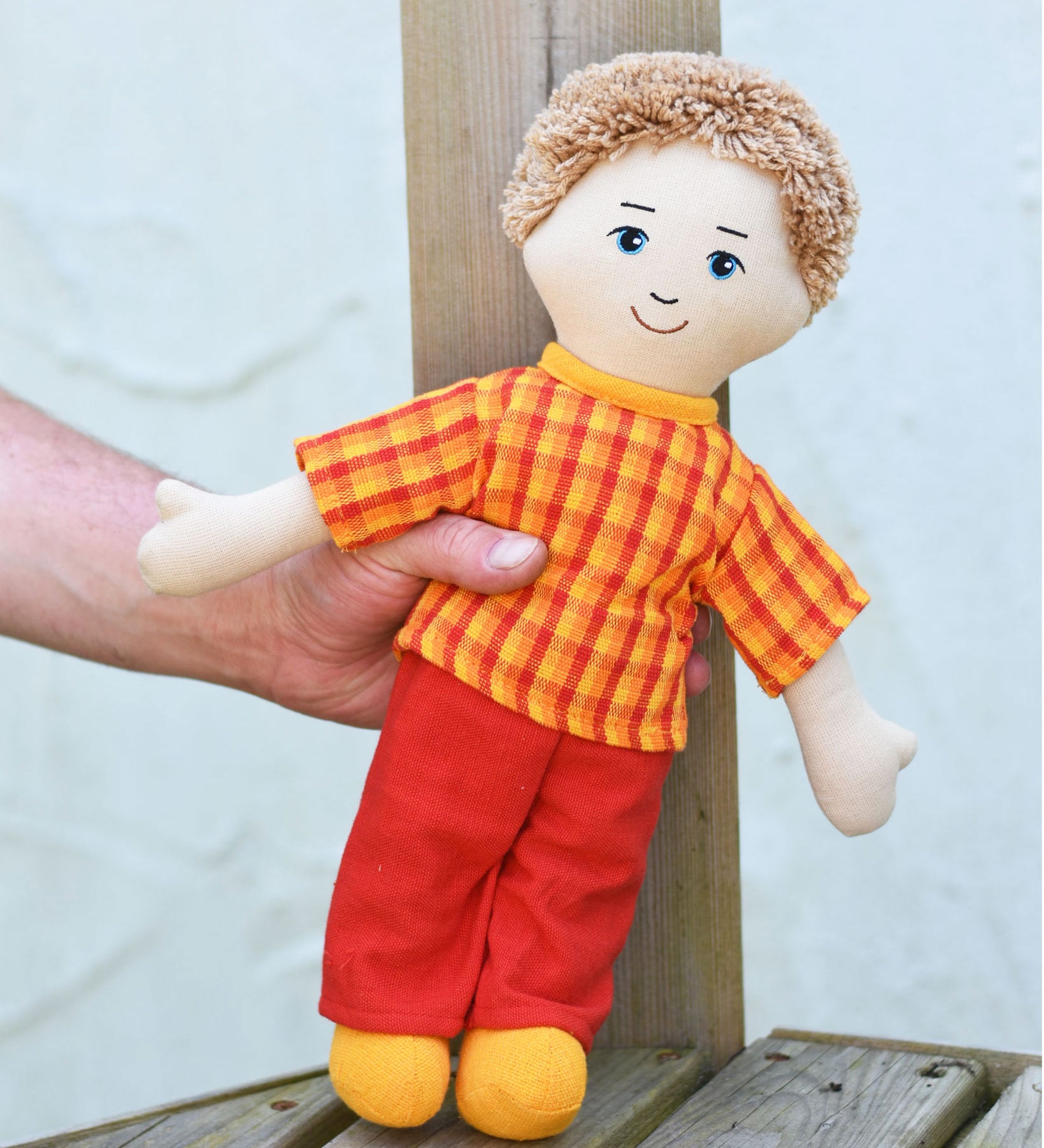 A Fair Trade Lanka Kade Dad doll shown in an adult's hand. The doll has white skin and white hair and is wearing a t-shirt and trousers made from sari material offcuts. 