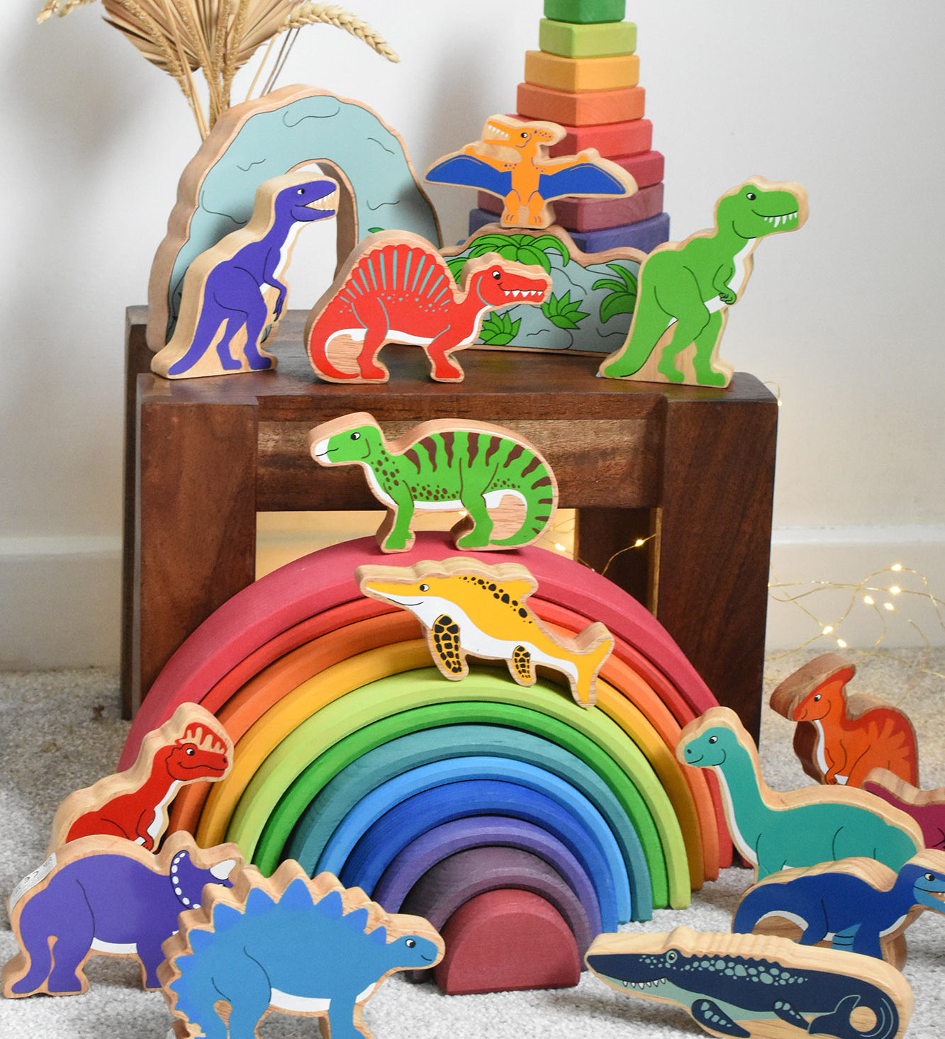 A large collection of Lanka Kade wooden toy dinosaur figures placed on a small wooden table and a Grimm's rainbow. These wooden toy figures are part of a huge range of Lanka Kade fair trade, wooden toys available here at Babipur.