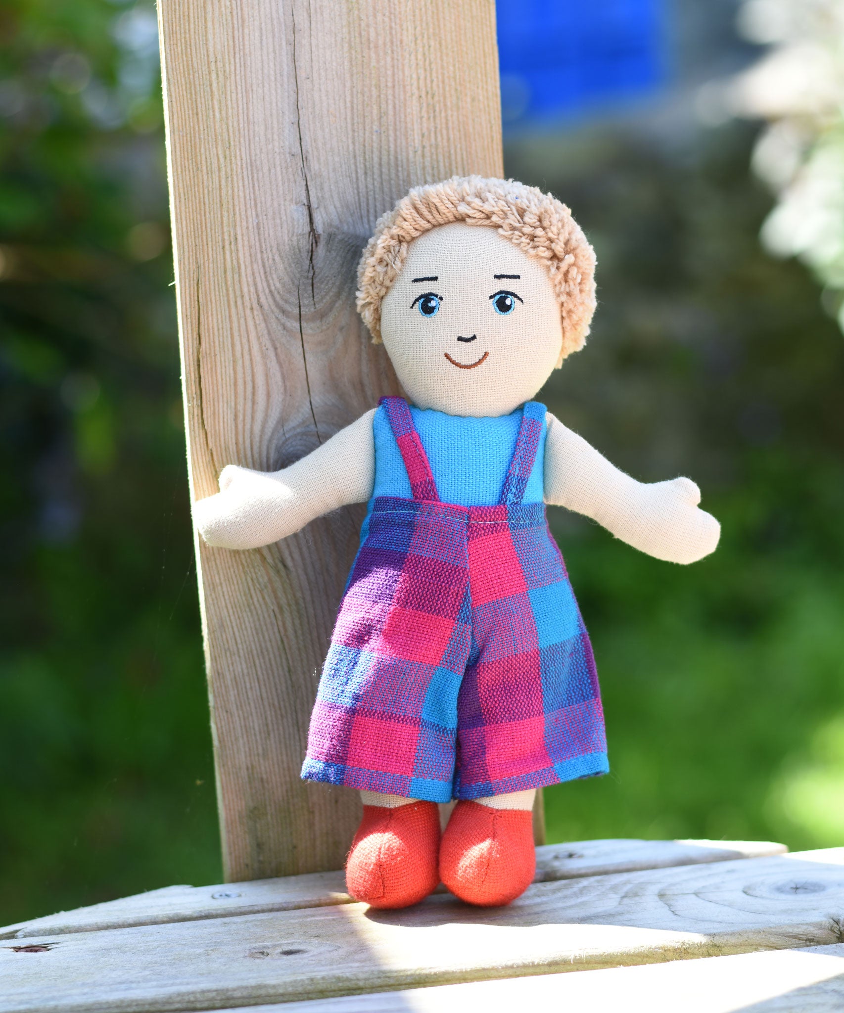 A Lanka Kade Boy Doll with White Skin and Blonde Hair leaning against a wooden fence post.