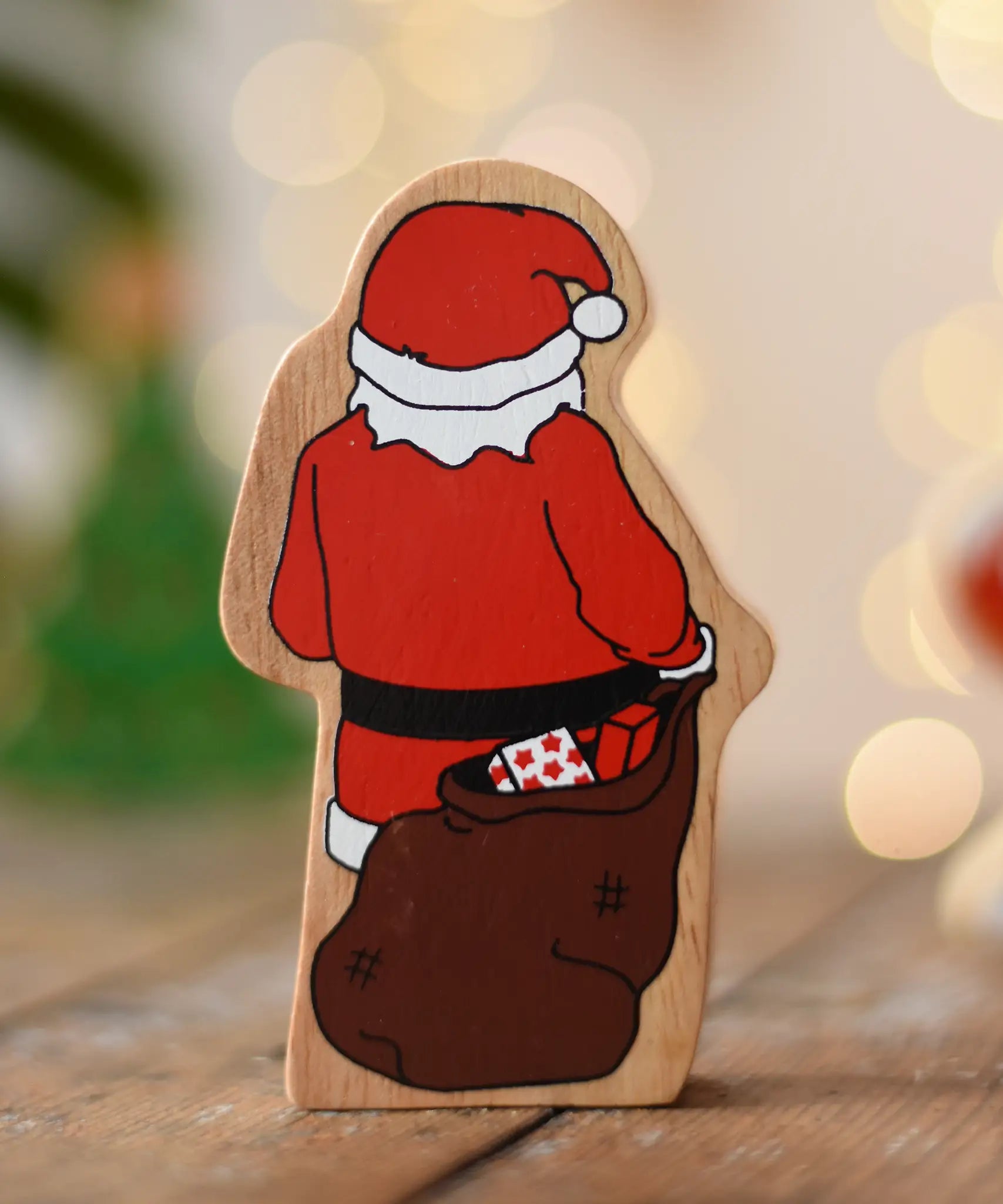 Back of the Lanka Kade Father Christmas wooden wooden toy figure placed on a wooden surface in the Babipur playroom. Santa is holding a brown sack filled with wrapped gifts.