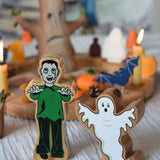 A Lanka Kade green and black Frankenstein style monster wooden figure and ghost figure placed in front of a Halloween themed Grimm's celebration ring set up. 