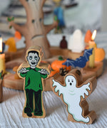 A Lanka Kade green and black Frankenstein style monster wooden figure and ghost figure placed in front of a Halloween themed Grimm's celebration ring set up. 