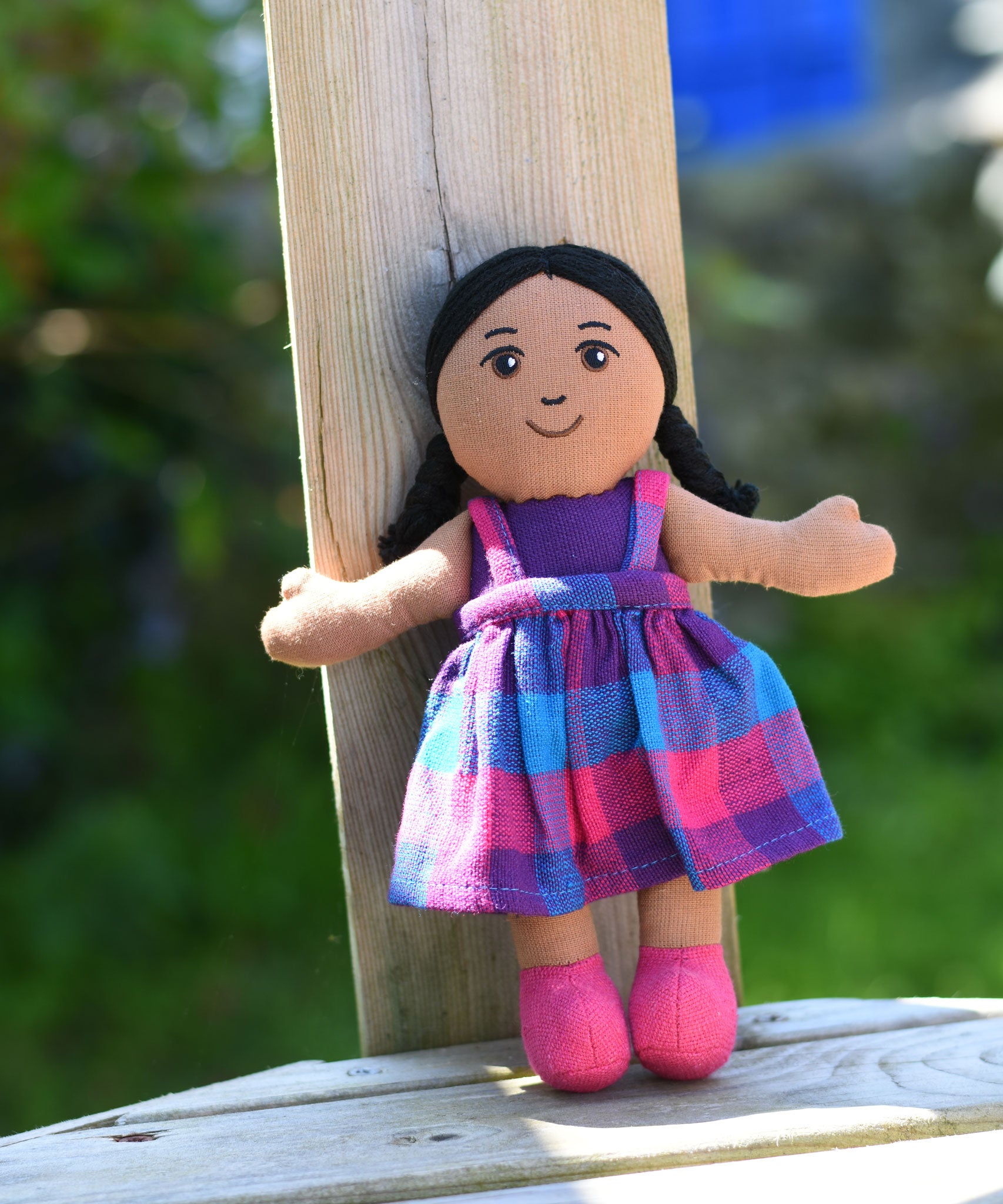 A Lanka Kade Girl Doll with Brown Skin and Black Hair leaning against a wooden post outdoors.