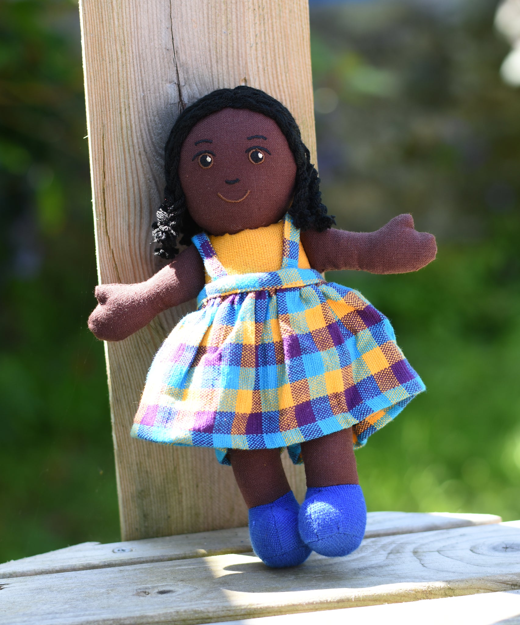 A Lanka Kade Girl Doll with Black Skin and Black Hair leaning against a wooden post outdoors.