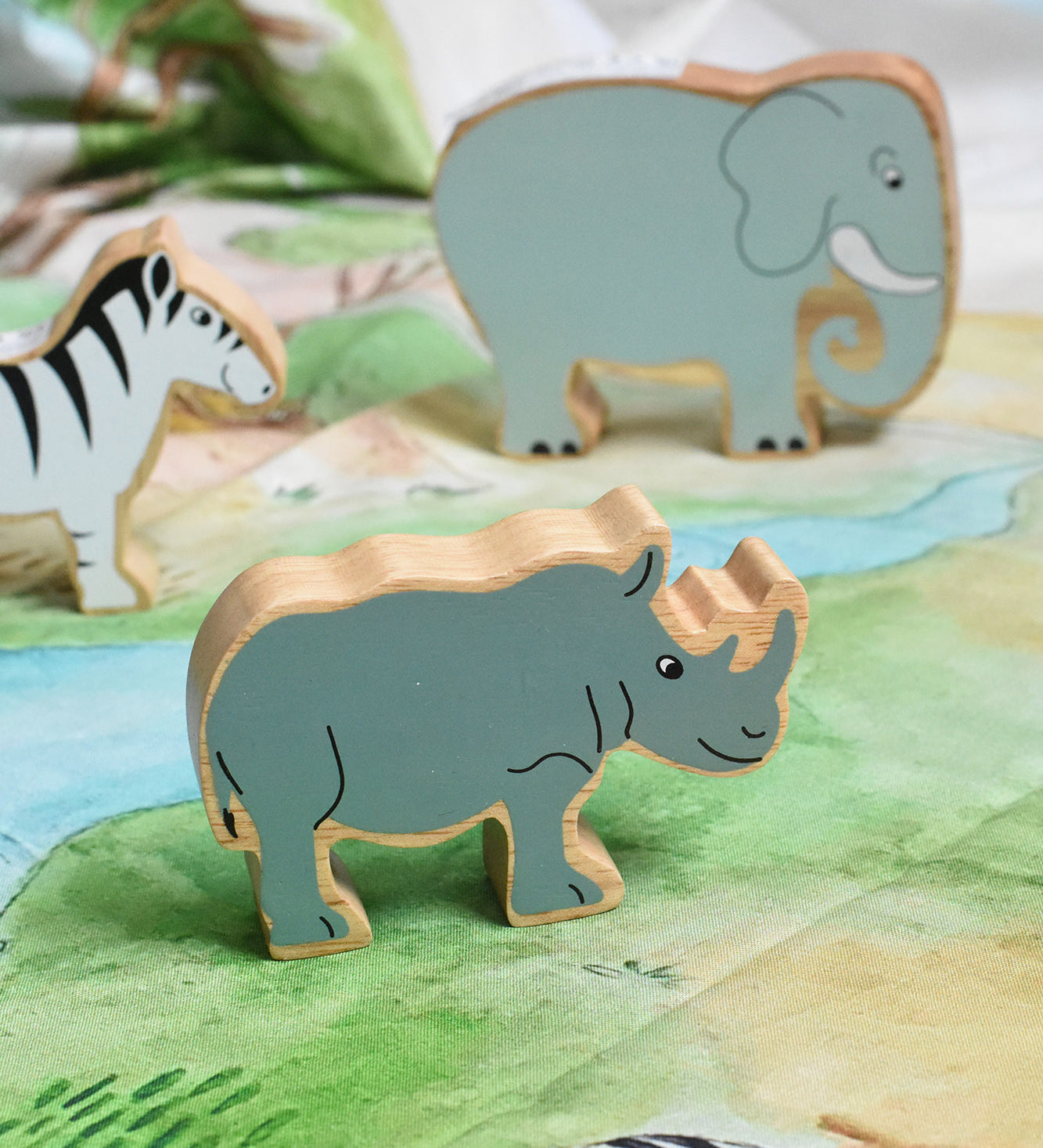 A Lanka Kade Grey Rhino Wooden Wild Animal Figure placed on a Wonderie Plains of Africa play cloth. An elephant and zebra figure can be seen in the background. 
These wooden toy figures are part of a huge range of Lanka Kade fair trade, wooden toys available here at Babipur.