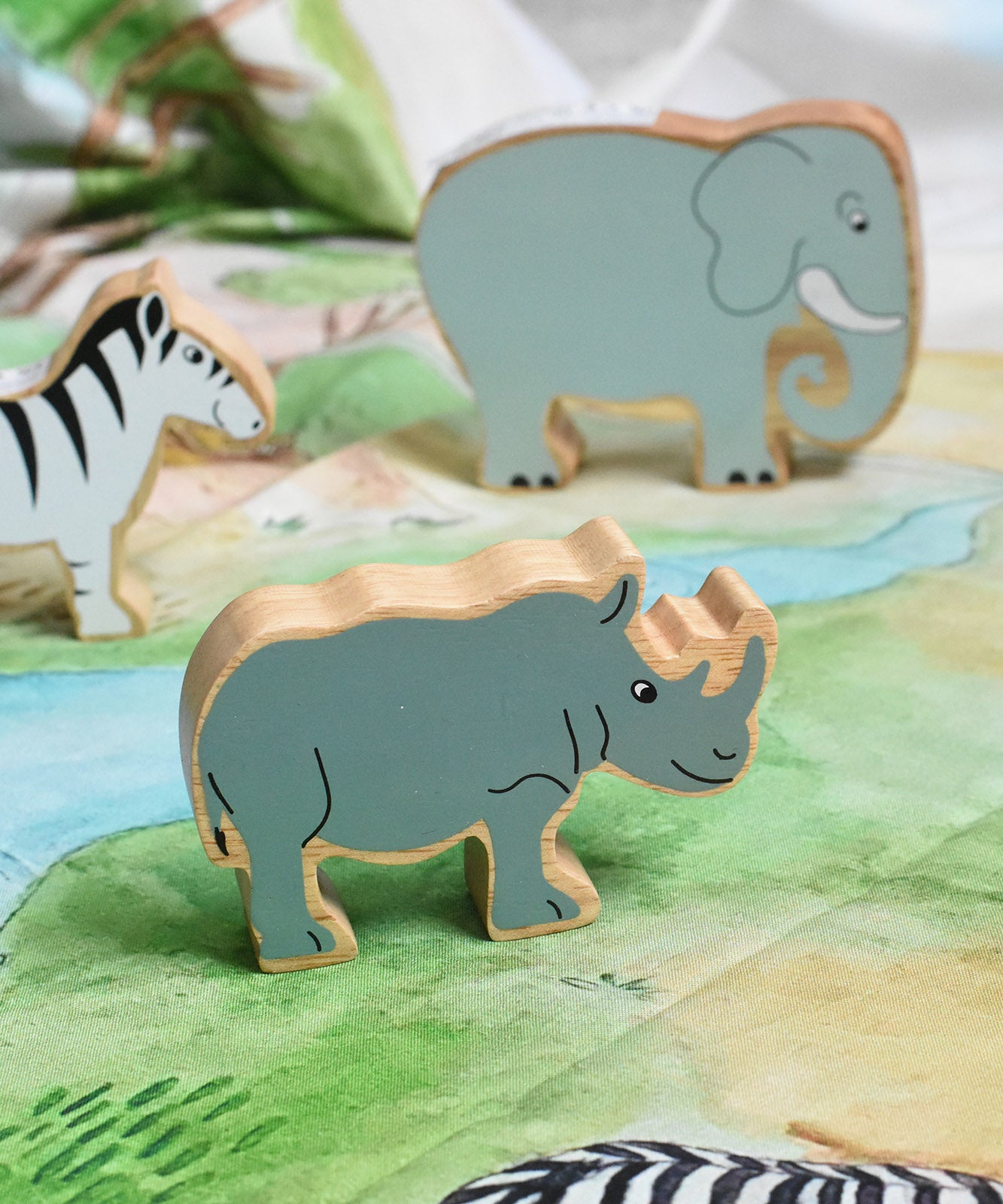 A Lanka Kade Grey Rhino Wooden Wild Animal Figure placed on a Wonderie Plains of Africa play cloth. An elephant and zebra figure can be seen in the background. 
These wooden toy figures are part of a huge range of Lanka Kade fair trade, wooden toys available here at Babipur.