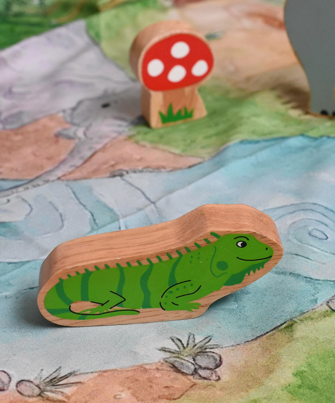 A Lanka Kade green iguana wooden toy figure placed on a Wonderie play cloth with a Babipur Lanka Kade mushroom in the background. These wooden toy figures are part of a huge range of Lanka Kade fair trade, wooden toys available here at Babipur.