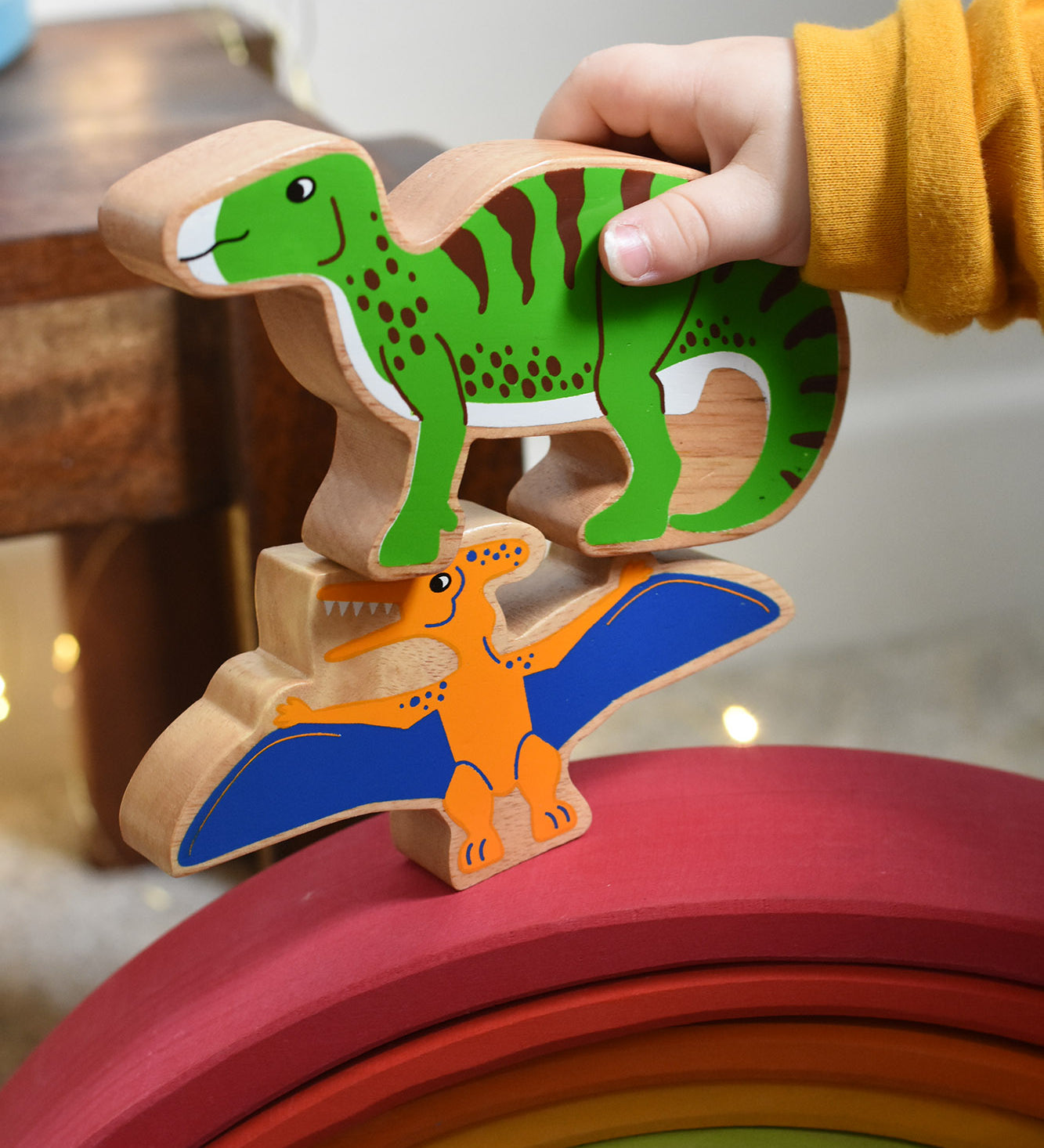 A close up of a child's hand holding the Lanka Kade green iguanodon wooden toy figure, placing it on a pteranodon figure. These wooden toy figures are part of a huge range of Lanka Kade fair trade, wooden toys available here at Babipur.