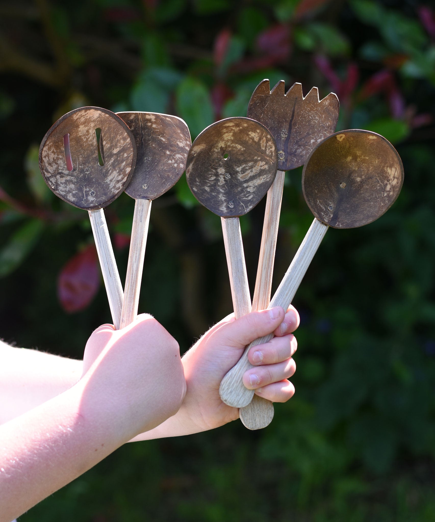 A child holding up the Lanka Kade Sri Lankan Coconut Shell Kitchen Utensils Playset in their hands. The set consists of 5 different coconut shell utensils on wooden handles.