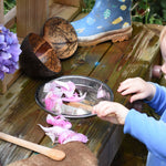A child playing with a coconut shell utensil set, scooping pink peony petals from a bowl of water. 