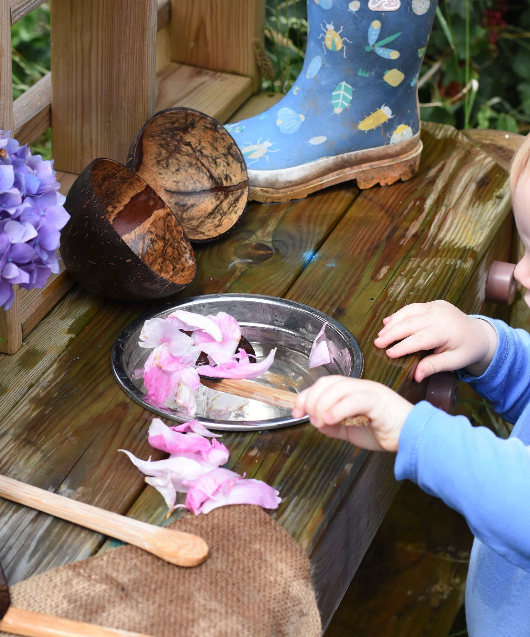 A child playing with a coconut shell utensil set, scooping pink peony petals from a bowl of water. 