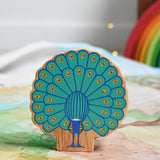 The back of a Lanka Kade peacock wooden toy figure placed on a Wonderie play cloth. The peacock toy has it's feather on display. These wooden toy figures are part of a huge range of Lanka Kade fair trade, wooden toys available here at Babipur.