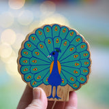 A Lanka Kade peacock wooden toy figure in an adult's hand. The peacock toy has it's feather on display. These wooden toy figures are part of a huge range of Lanka Kade fair trade, wooden toys available here at Babipur.