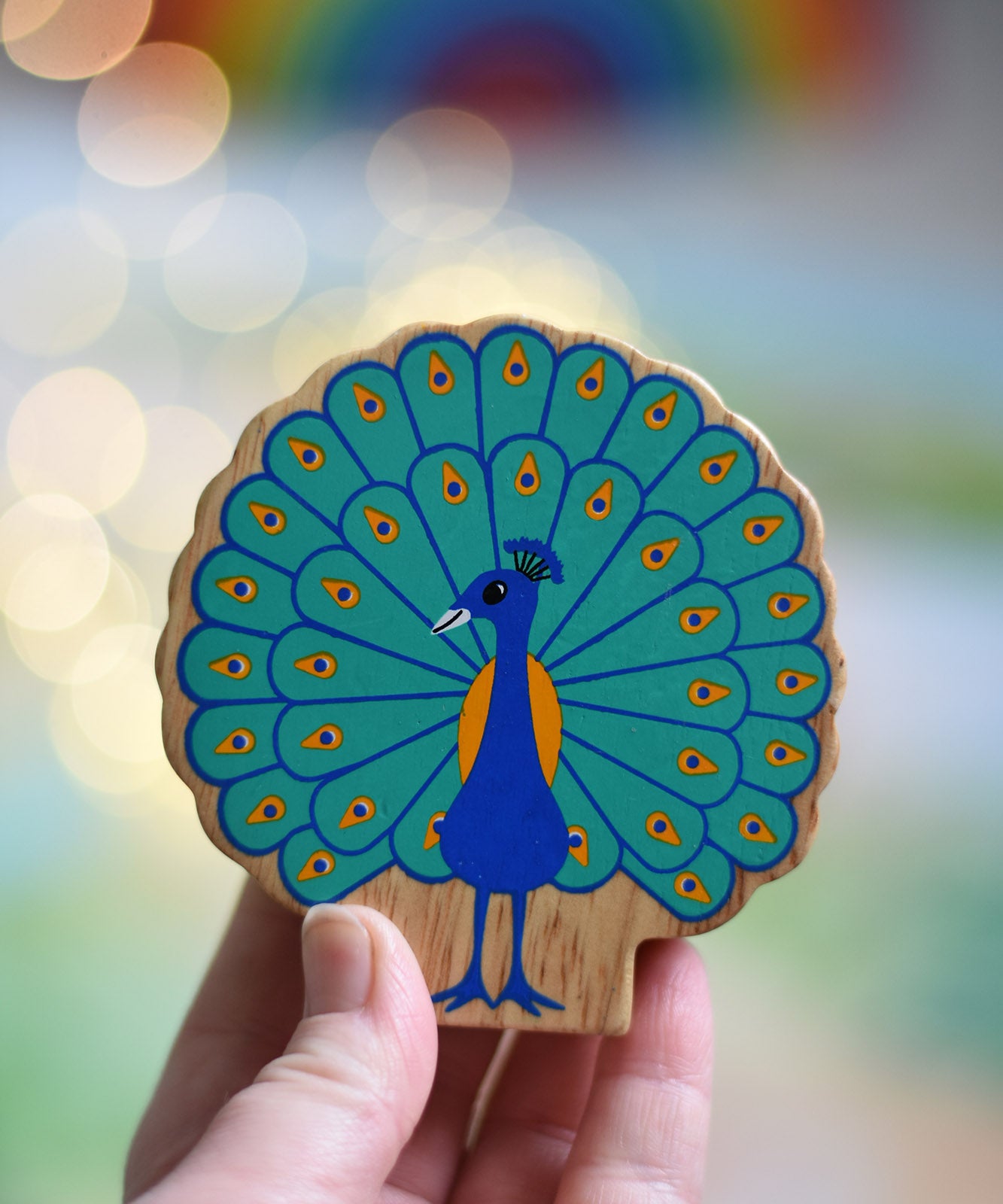 A Lanka Kade peacock wooden toy figure in an adult's hand. The peacock toy has it's feather on display. These wooden toy figures are part of a huge range of Lanka Kade fair trade, wooden toys available here at Babipur.