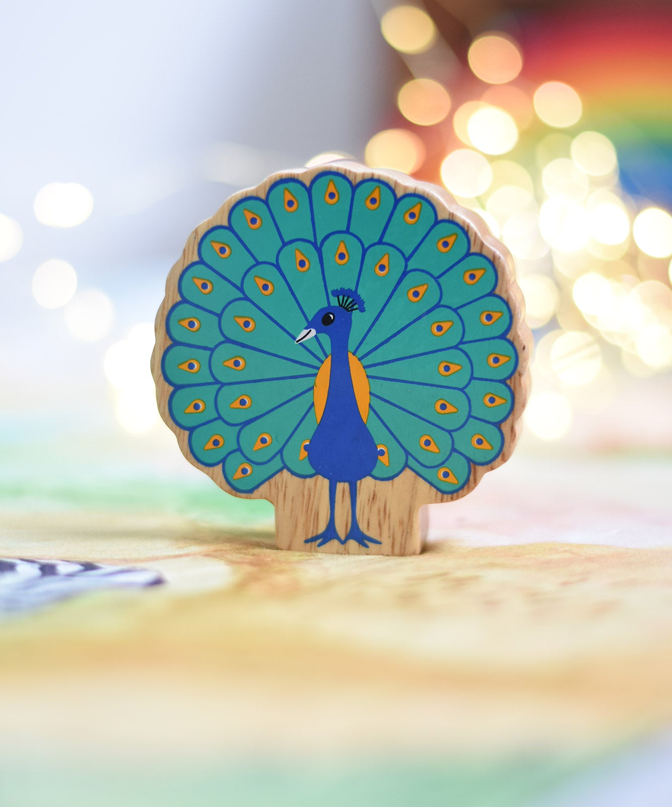 A Lanka Kade peacock wooden toy figure placed on a Wonderie play cloth. The peacock toy has it's feather on display. These wooden toy figures are part of a huge range of Lanka Kade fair trade, wooden toys available here at Babipur.