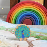 A Lanka Kade peacock wooden toy figure placed on a Wonderie play cloth. The peacock toy has it's feather on display. A Grimm's rainbow can be seen in the background. These wooden toy figures are part of a huge range of Lanka Kade fair trade, wooden toys available here at Babipur.