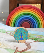 A Lanka Kade peacock wooden toy figure placed on a Wonderie play cloth. The peacock toy has it's feather on display. A Grimm's rainbow can be seen in the background. These wooden toy figures are part of a huge range of Lanka Kade fair trade, wooden toys available here at Babipur.
