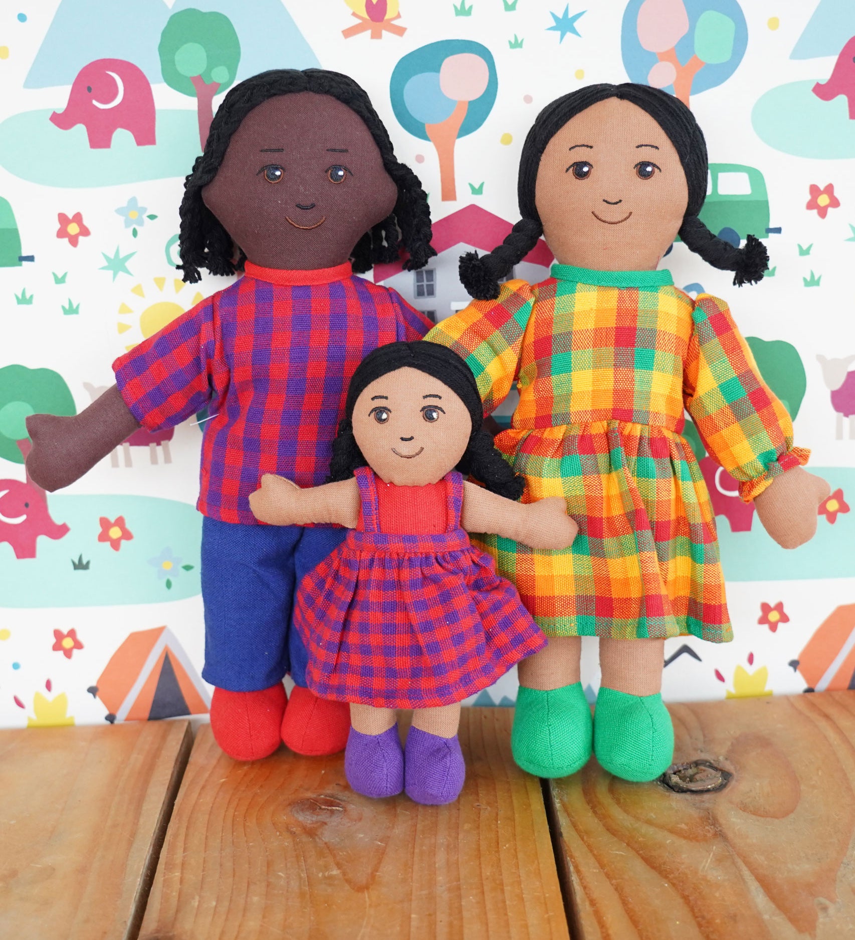 A Lanka Kade Fair Trade Rag Doll Dad with black Skin and black hair, Mum with brown skin and black hair and daughter with brown skin and black hair. The rag dolls are in a standing position on a wooden surface. 