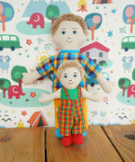A Lanka Kade Fair Trade Rag Doll Dad with White Skin and Blonde hair with a son rag doll. The dad is in a standing position leaning against a patterned wallpapered wall with the son doll standing in front of him. 