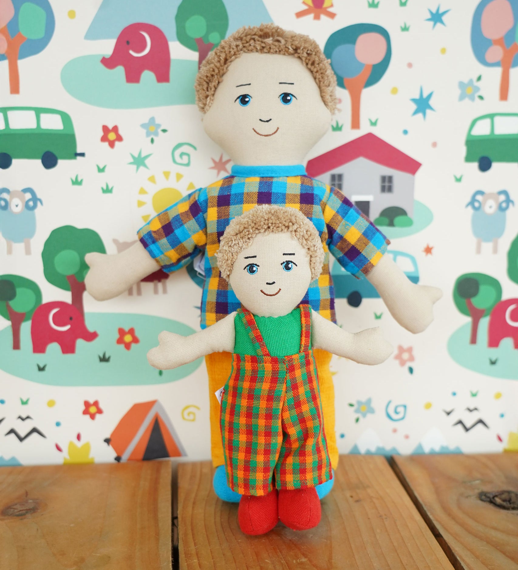 A Lanka Kade Fair Trade Rag Doll Dad with White Skin and Blonde hair with a son rag doll. The dad is in a standing position leaning against a patterned wallpapered wall with the son doll standing in front of him. 