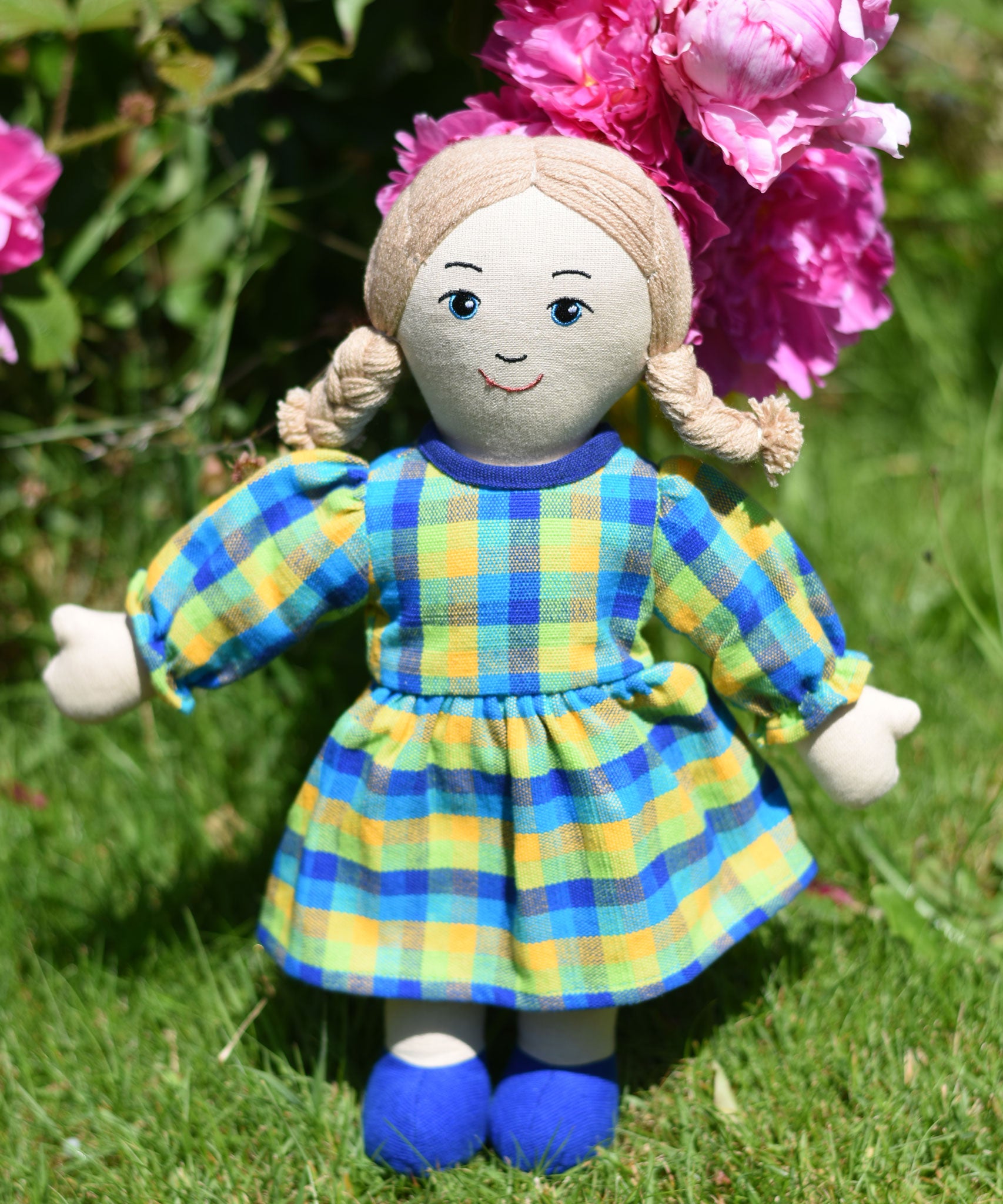 A Lanka Kade Rag Doll Mum with White Skin and Blonde Hair placed on grass in front of peonies growing in a garden.
