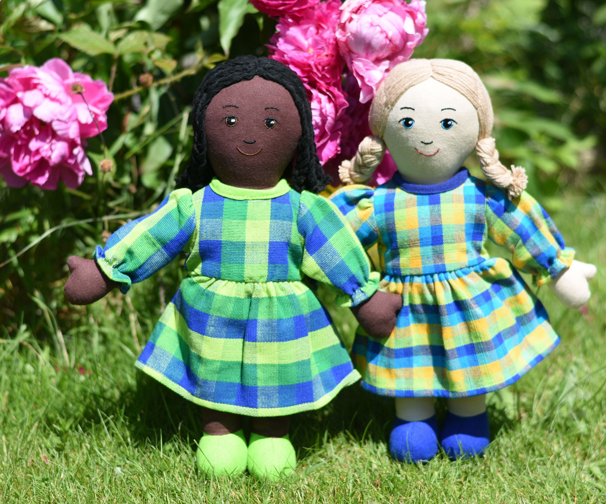 Two Lanka Kade Rag Doll Mums one with black hair and black skin White Skin, Blonde Hair placed on grass in front of peonies growing in a garden.