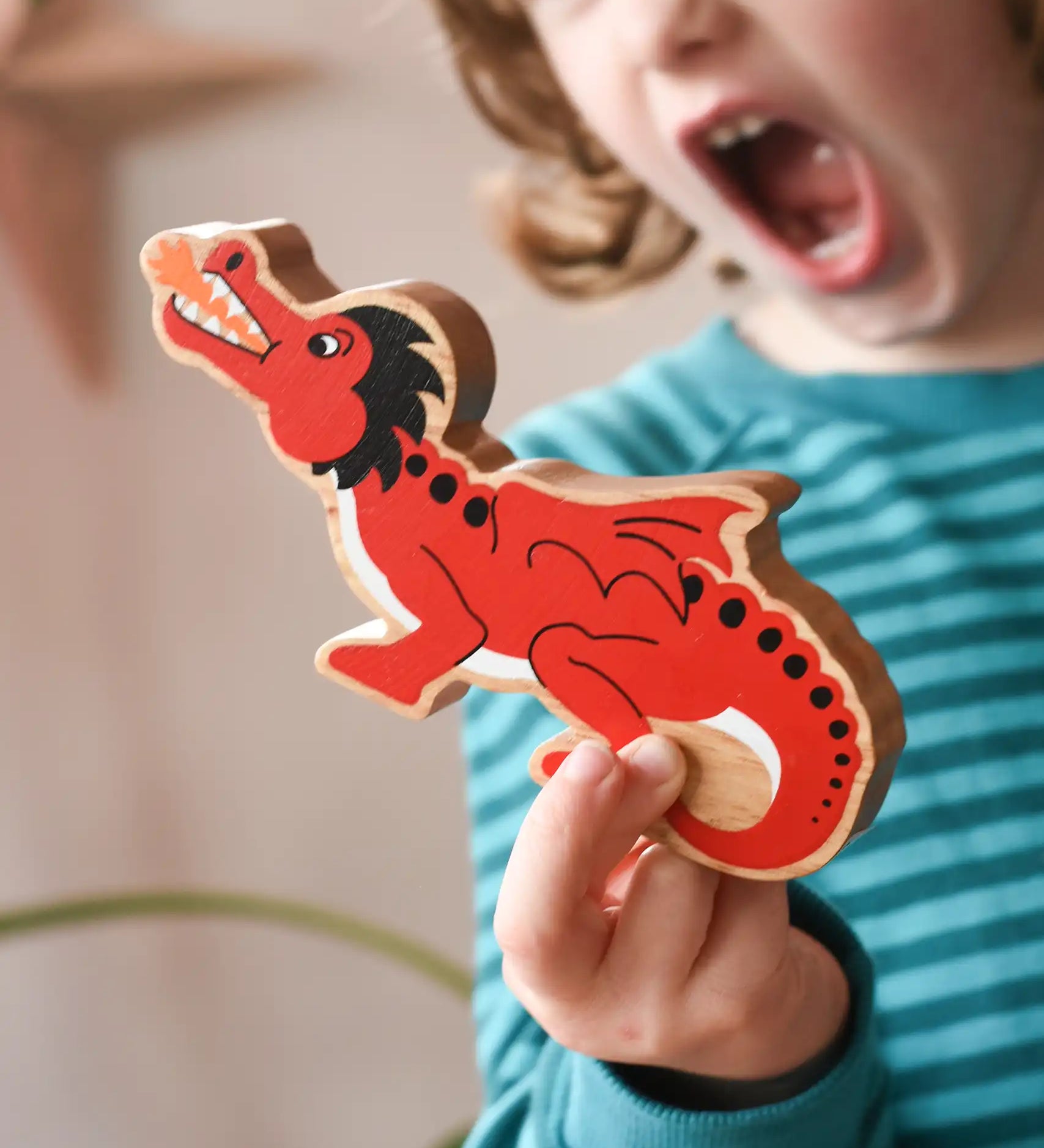 Lanka Kade wooden toy dragon figure in red with black, white and orange details in a child's hand. The child can be seen roaring in the background.