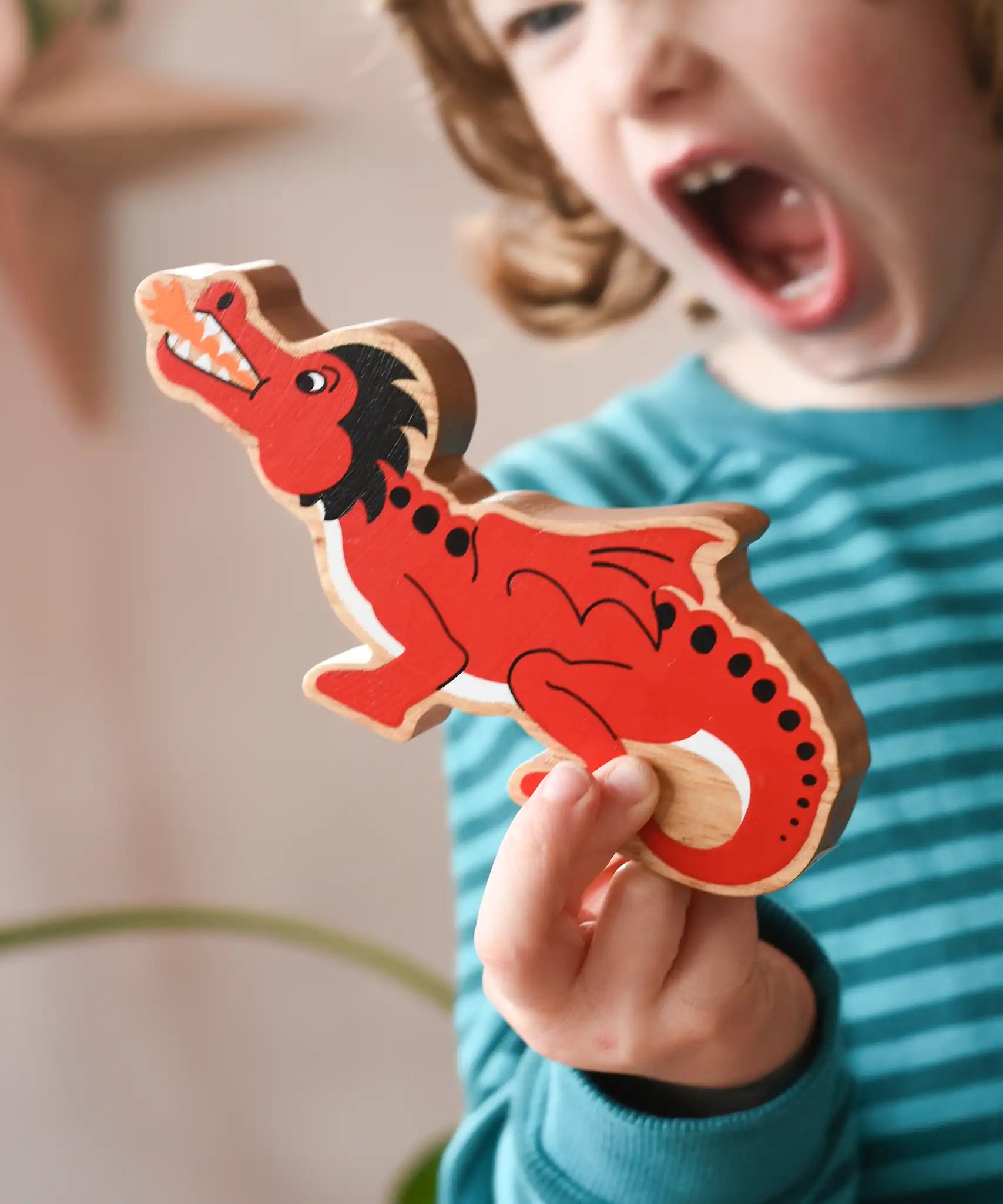 Lanka Kade wooden toy dragon figure in red with black, white and orange details in a child's hand. The child can be seen roaring in the background.