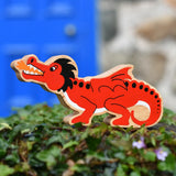 Lanka Kade red dragon wooden toy dragon figure with black details, standing on green foliage in front of a blurred blue door and stone wall.
