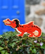 Lanka Kade red dragon wooden toy dragon figure with black details, standing on green foliage in front of a blurred blue door and stone wall.