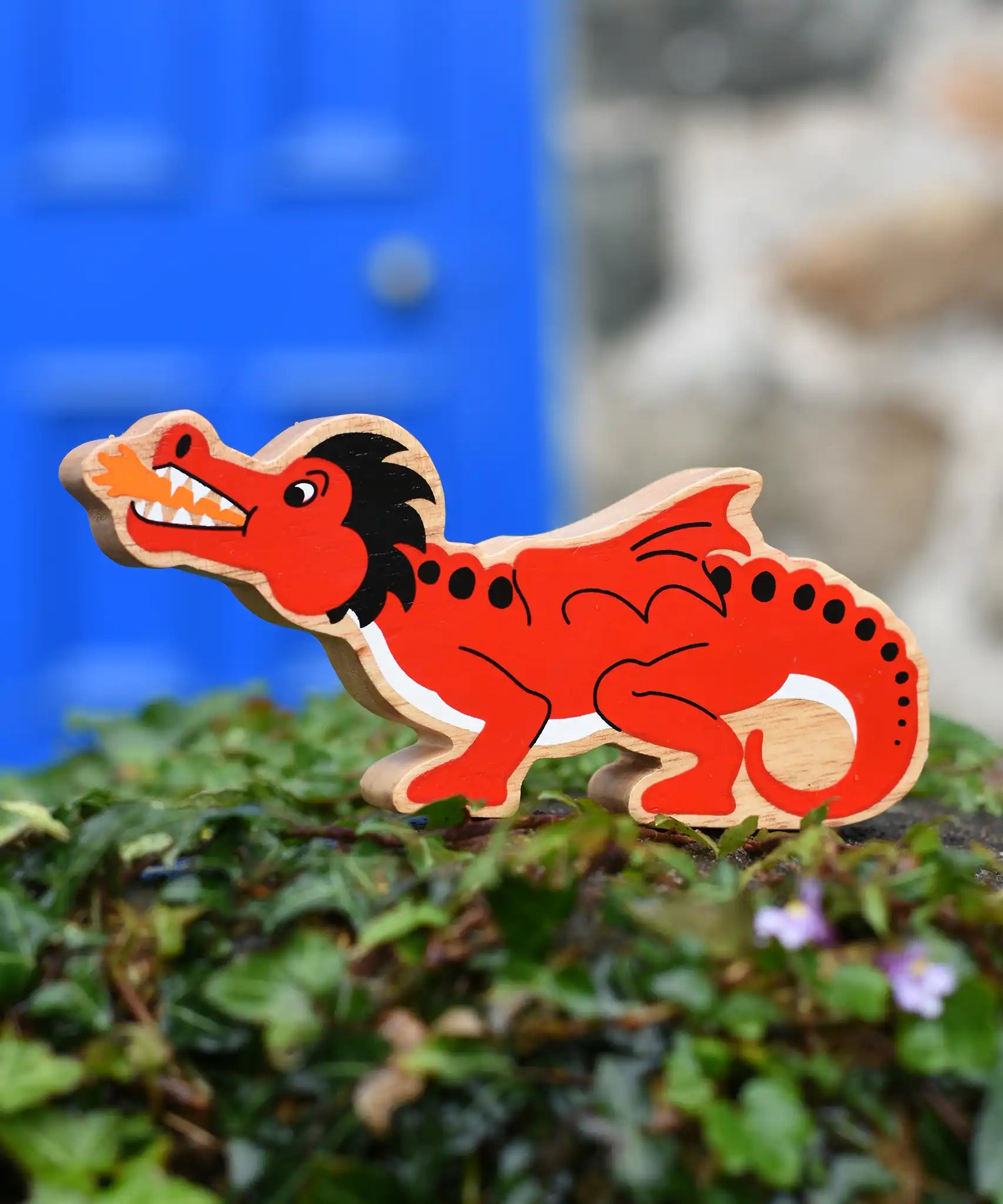 Lanka Kade red dragon wooden toy dragon figure with black details, standing on green foliage in front of a blurred blue door and stone wall.
