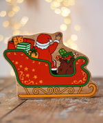 Back of the Lanka Kade Father Christmas in a sleigh wooden toy figure. The back features Santa sitting in the sleigh waving, surrounded by gifts. 