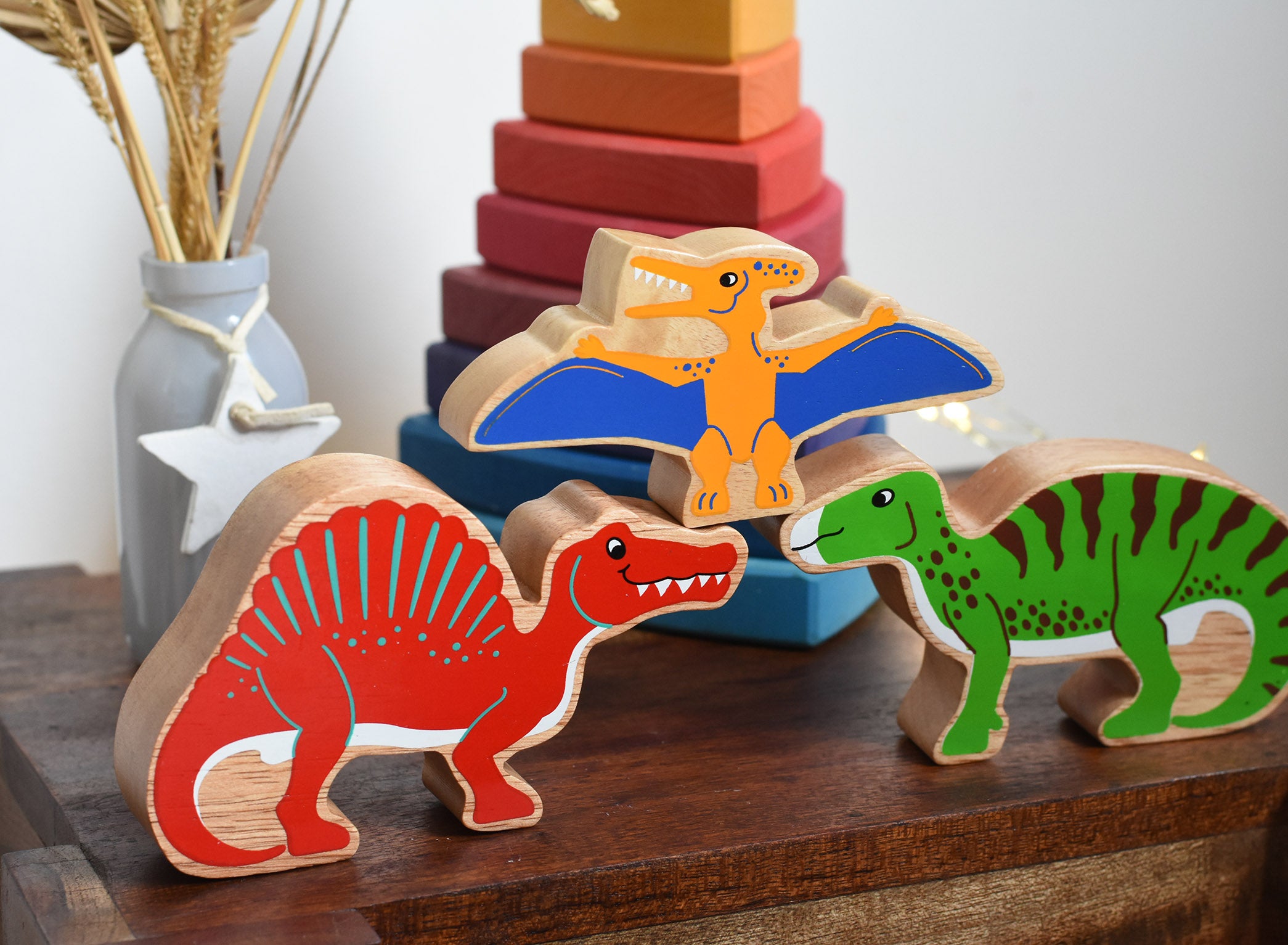 A collection of three Lanka Kade Wooden dinosaur figures placed on a small wooden table. A red Spinosaurus, a yellow pteranodon and a green iguanodon. These wooden toy figures are part of a huge range of Lanka Kade fair trade, wooden toys available here at Babipur.
