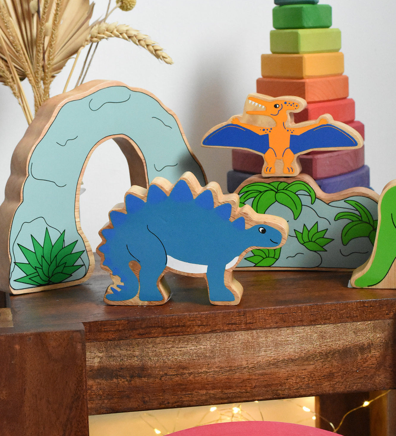 Lanka Kade Stegosaurus Dinosaur toy figure placed on a small wooden table with scenery pieces from the prehistoric play set in the background. These wooden toy figures are part of a huge range of Lanka Kade fair trade, wooden toys available here at Babipur.