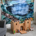 A Lanka Kade wooden toy castle placed on a carpeted floor in front of a Wonderie Moonlight Forest design play cloth that's been hung on the wall behind. Fairytale themed wooden toy character figures have been placed in and around the wooden toy castle. 