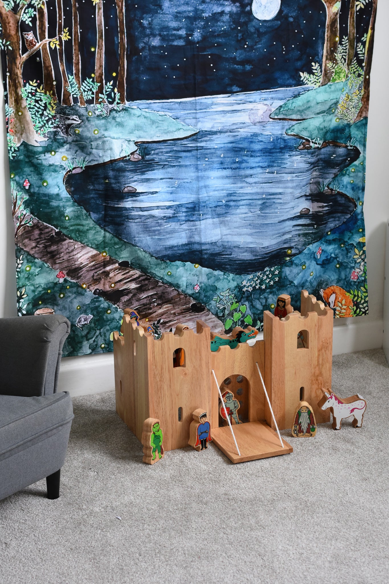 A Lanka Kade wooden toy castle placed on a carpeted floor in front of a Wonderie Moonlight Forest design play cloth that's been hung on the wall behind. Fairytale themed wooden toy character figures have been placed in and around the wooden toy castle. 