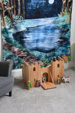 A Lanka Kade wooden toy castle placed on a carpeted floor in front of a Wonderie Moonlight Forest design play cloth that's been hung on the wall behind. Fairytale themed wooden toy character figures have been placed in and around the wooden toy castle. 