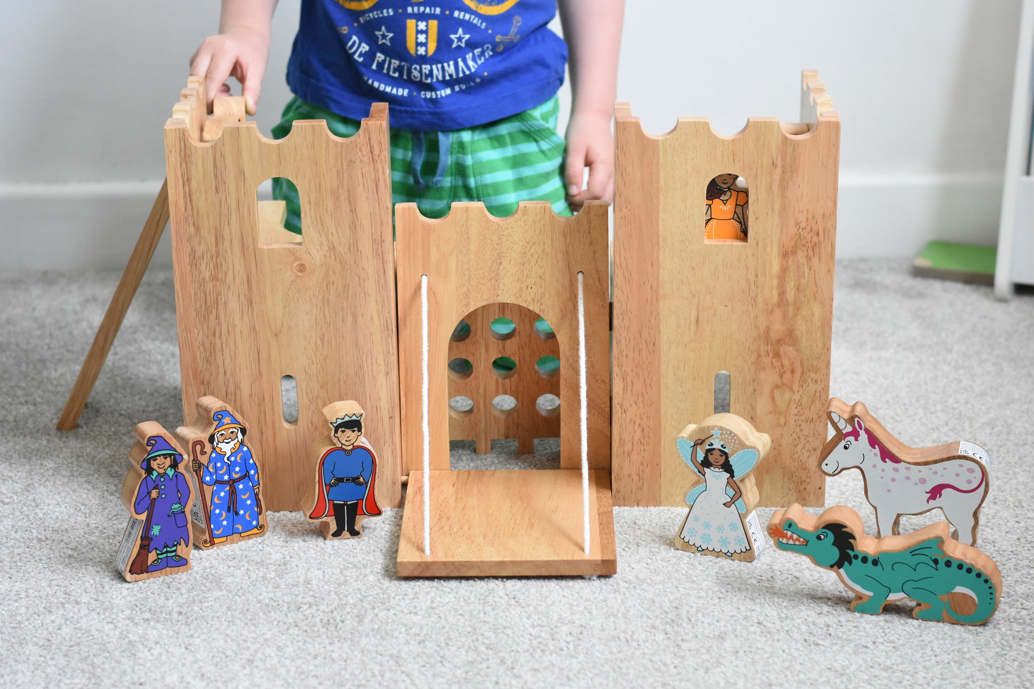 The Lanka Kade wooden toy fairytale castle play set shown with the wooden character figures in and around the castle. A witch, wizard, prince, fairy, unicorn and dragon can be seen standing in front of the castle, whilst a princess can be seen peering through one of the windows of the castle. 