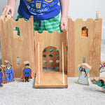 The Lanka Kade wooden toy fairytale castle play set shown with the wooden character figures in and around the castle. A witch, wizard, prince, fairy, unicorn and dragon can be seen standing in front of the castle, whilst a princess can be seen peering through one of the windows of the castle. 