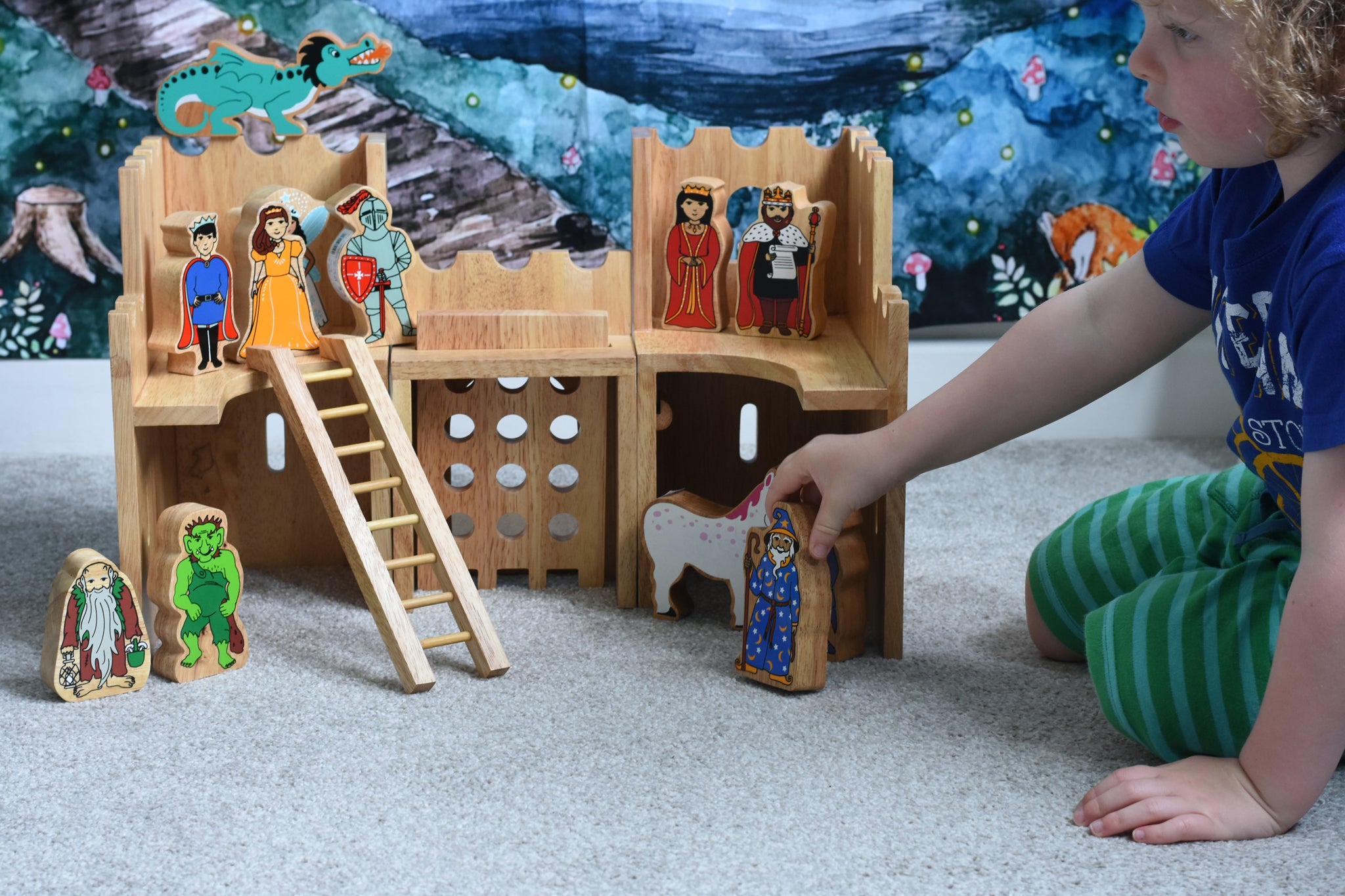 A child playing with the Lanka Kade Fairytale castle playset. The fairytale themed wooden character figures have been placed in and around the toy castle.  The view is of the back or inside of the castle. A moveable toy ladder is included in the set. 