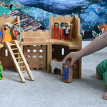 A child playing with the Lanka Kade Fairytale castle playset. The fairytale themed wooden character figures have been placed in and around the toy castle.  The view is of the back or inside of the castle. A moveable toy ladder is included in the set. 