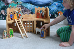 A child playing with the Lanka Kade Fairytale castle playset. The fairytale themed wooden character figures have been placed in and around the toy castle.  The view is of the back or inside of the castle. A moveable toy ladder is included in the set. 