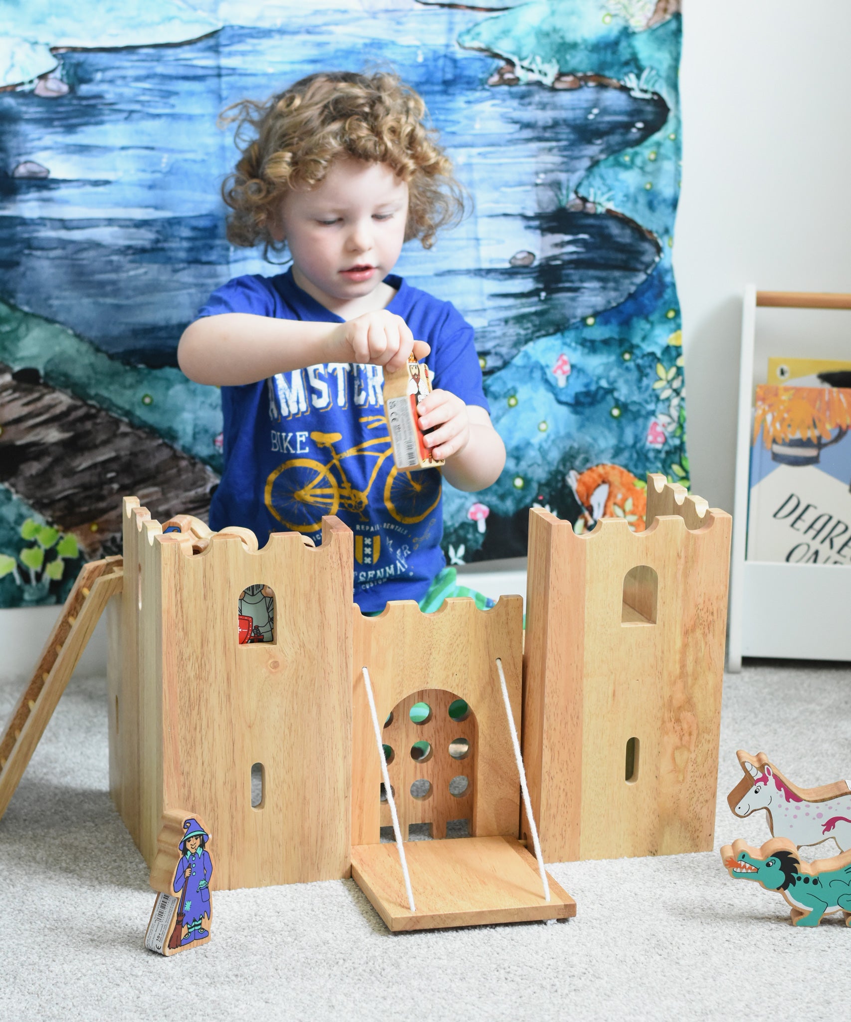 A child playing with the Lanka Kade wooden toy fairytale castle play set. A Wonderie moonlight forest can be seen hung on the wall in the background. 